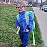 child, boy, glasses, backpack, blue_clothing, nike, grass, sidewalk, brick_building, car, outdoor, casual, walking, smiling, daylight, urban, person, trees, jacket, pants