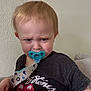 toddler, child, pacifier, grumpy, rolling_stones, tshirt, indoor, couch, blond_hair, blue_eyes, expression, person, seated, red_blanket, casual_clothing, home, young_child, face, portrait, baby