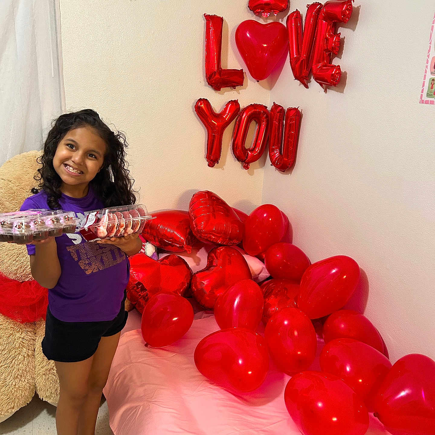 Viviana is registered to the contest to win money with this photo: child, girl, smile, cupcakes, balloons, heart_shaped, red, bed, pink_bedspread, barefoot, curly_hair, indoor, celebration, love, wall, decorations, holding, food, happy, person