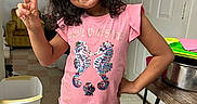 Viviana is registered to the contest to win money with this photo: child, girl, smiling, indoor, peace_sign, pink_shirt, seahorses, plaid_pants, curly_hair, trash_bin, table, poster, batman, door, floor, household, person, happy, casual, standing