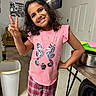 child, girl, smiling, indoor, peace_sign, pink_shirt, seahorses, plaid_pants, curly_hair, trash_bin, table, poster, batman, door, floor, household, person, happy, casual, standing
