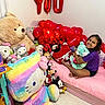 Viviana is registered to the contest to win money with this photo: child, smiling, plush_toy, stuffed_animal, balloon, heart_shaped, bed, pink_bedspread, room, happy, toy, colorful, decor, wall, indoor, cuddly, celebration, cozy, person, sitting