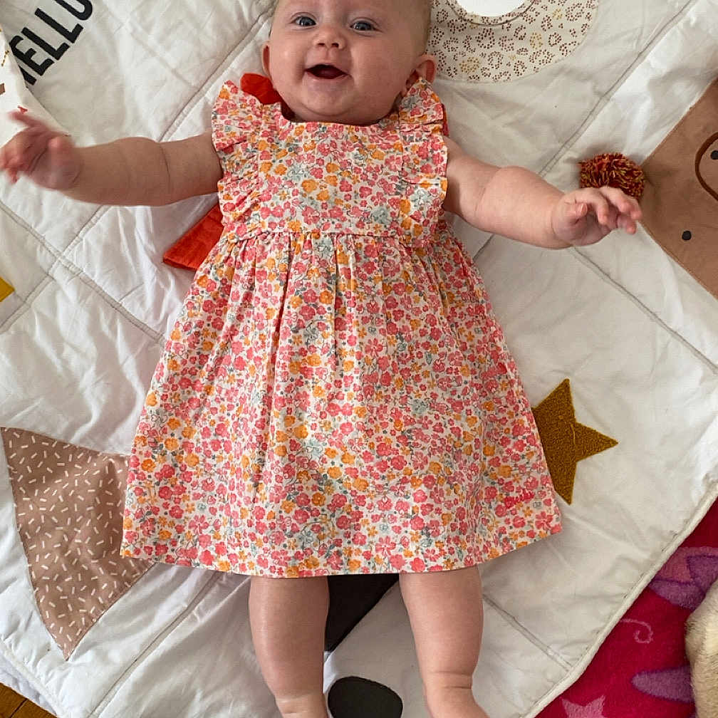 Charlee a rejoint le concours — aidez-le/la à gagner de superbes lots ! arms_outstretched, baby, cheerful, child, colorful, cute, feet, floor, floral_dress, happy, indoors, infant, lying_down, panda_design, patterned_mat, play_mat, portrait, smiling, soft_toys, wooden_floor