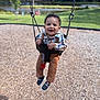 Jamir is registered to the contest to win money with this photo: toddler, child, swing, playground, smiling, happy, outdoor, park, grass, pond, trees, plaid_shirt, corduroy_pants, black_shoes, pacifier, sunny, daylight, person, fun, nature