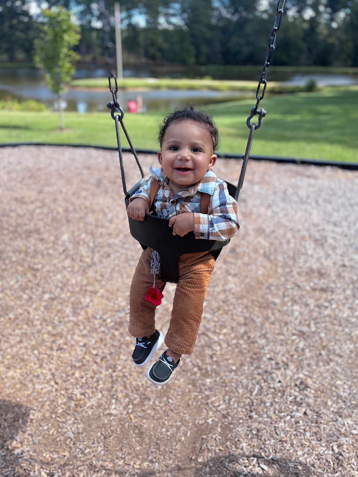 Jamir is registered to the contest to win money with this photo: toddler, child, swing, playground, smiling, happy, outdoor, park, grass, pond, trees, plaid_shirt, corduroy_pants, black_shoes, pacifier, sunny, daylight, person, fun, nature
