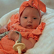 Clara is registered to the contest to win money with this photo: baby, infant, orange_outfit, headband, pacifier, blanket, soft_texture, lying_down, cute, child, newborn, comfort, portrait, indoors, person, apparel, sleepwear, resting, young_child, cozy