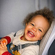 Fabio a rejoint le concours — aidez-le/la à gagner de superbes lots ! child, toddler, smiling, high_chair, orange_toy, teeth, curly_hair, happy, portrait, indoor, cozy_clothing, harness, face, blue_eyes, hand, skin, fleece, closeup, grey_background, seat
