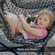 Skyler joined the competition — help win amazing prizes! child, cat, hammock_chair, outdoor, curly_hair, pink_shirt, pet, hugging, relaxing, concrete_floor, wooden_fence, grill, patterned_fabric, animal, person, comfort, daylight, young_child, resting, casual