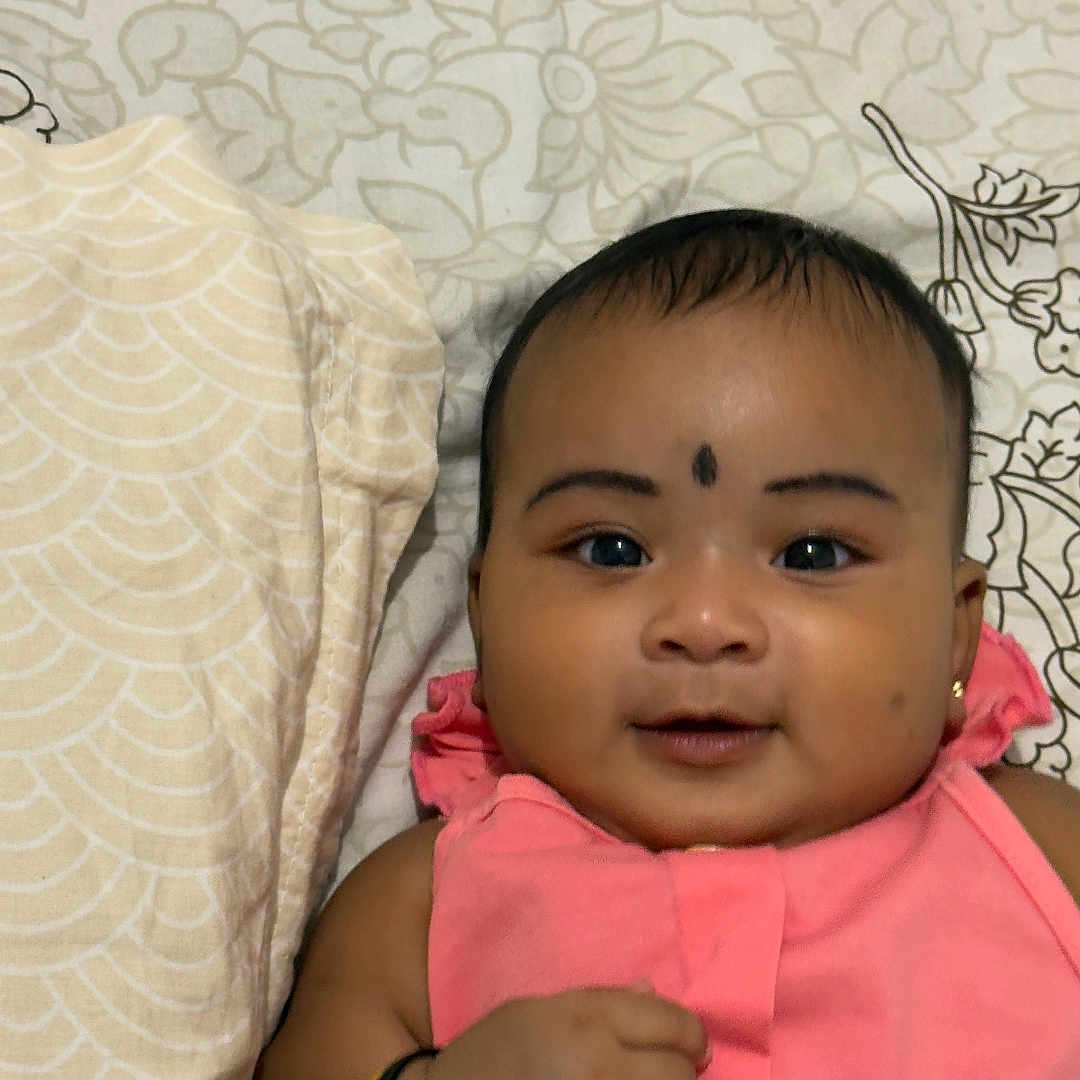 Ekaksha joined the competition — help win amazing prizes! adorable, baby, bedspread, bracelet, child, closeup, cute, face, hand, happy, indoors, infant, lying_down, patterned_background, person, pink_clothing, portrait, skin, smiling, young_child