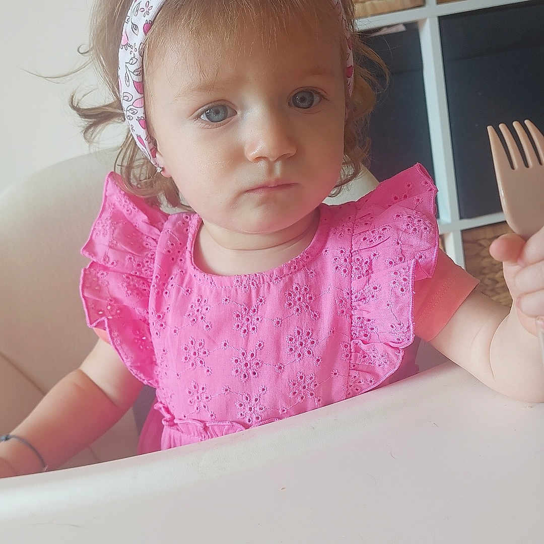 Juliette participe au concours pour gagner de l'argent avec cette photo : basket, blue_eyes, child, curly_hair, cute, feeding, fork, furniture, headband, high_chair, home, indoor, person, pink_dress, portrait, serious_expression, sitting, storage, table, toddler
