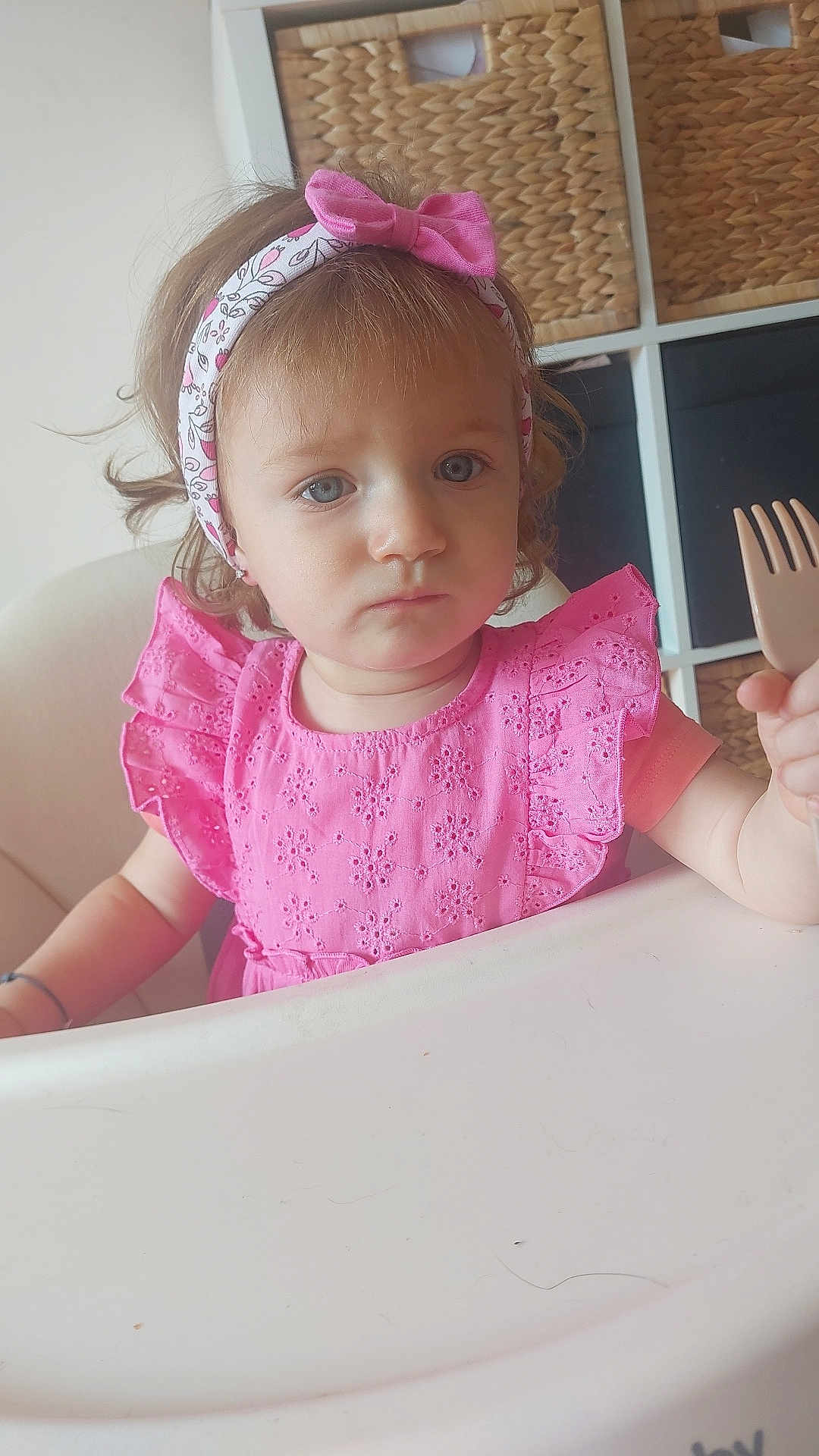 Juliette participe au concours pour gagner de l'argent avec cette photo : toddler, child, pink_dress, headband, fork, high_chair, indoor, basket, storage, furniture, curly_hair, blue_eyes, serious_expression, home, person, portrait, sitting, table, feeding, cute