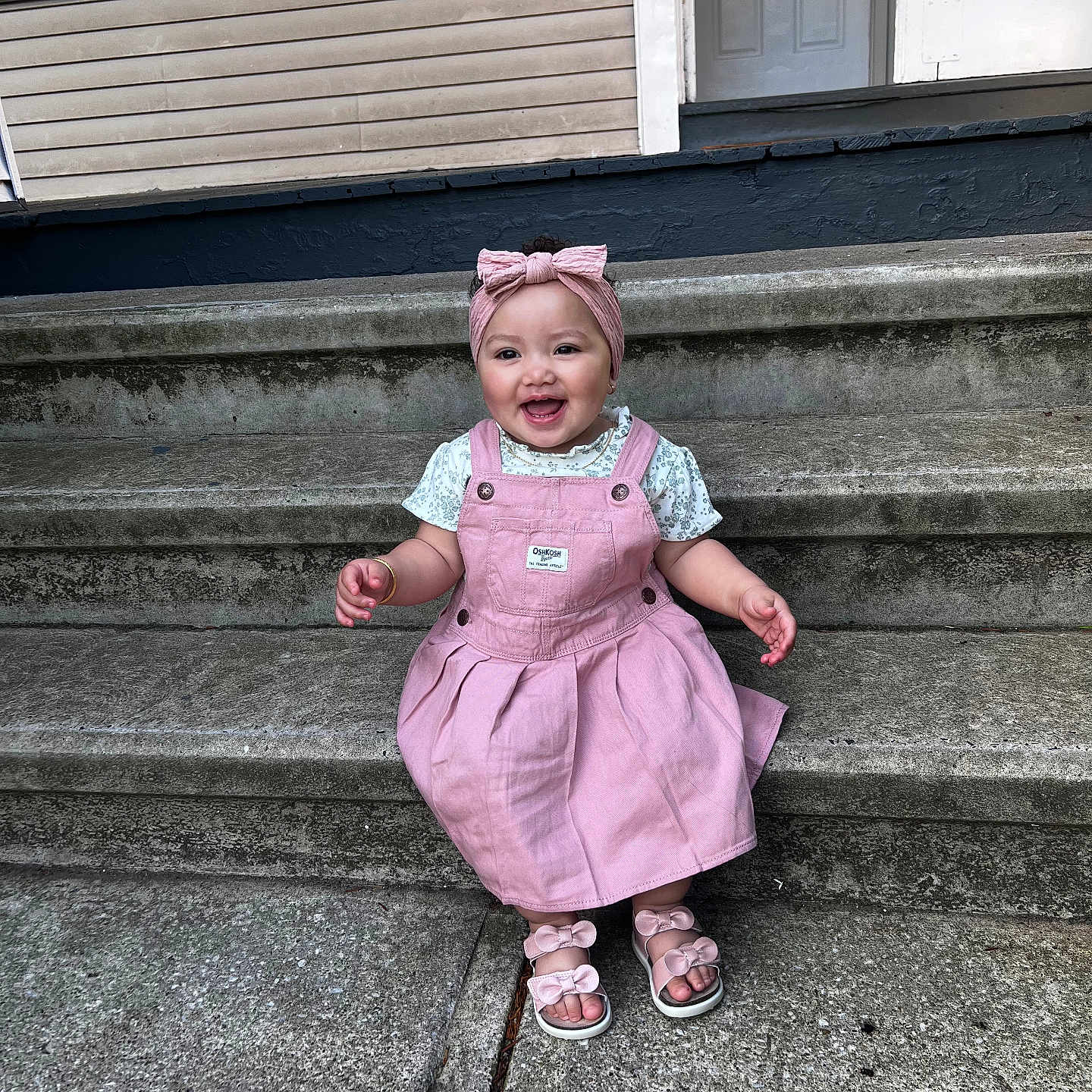 Malia is registered to the contest to win money with this photo: accessory, baby, child, concrete, cute, door, fashion, footwear, happy, headband, house, outdoor, person, pink_dress, portrait, sandals, smiling, steps, toddler, window