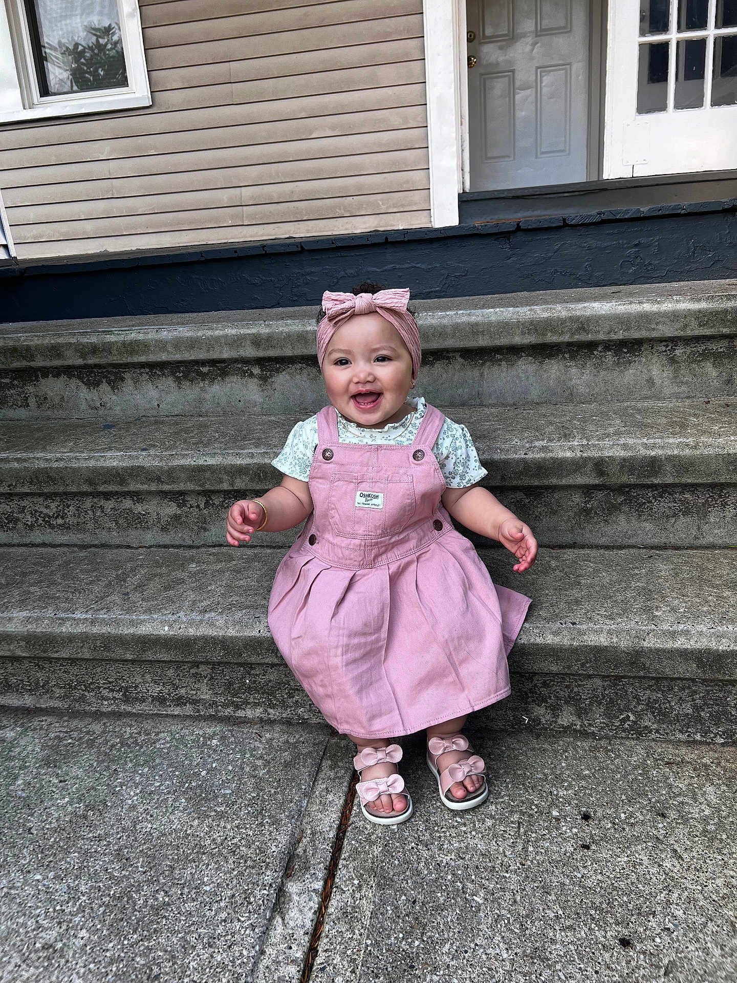 Malia is registered to the contest to win money with this photo: baby, child, smiling, pink_dress, headband, sandals, outdoor, steps, concrete, house, door, window, happy, cute, person, toddler, fashion, footwear, accessory, portrait
