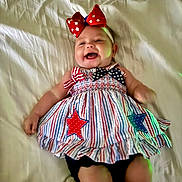 Kennedy is registered to the contest to win money with this photo: baby, child, smiling, lying_down, dress, headband, bow, stars, stripes, red, white, blue, fabric, bed, sheet, happy, cute, infant, person, portrait