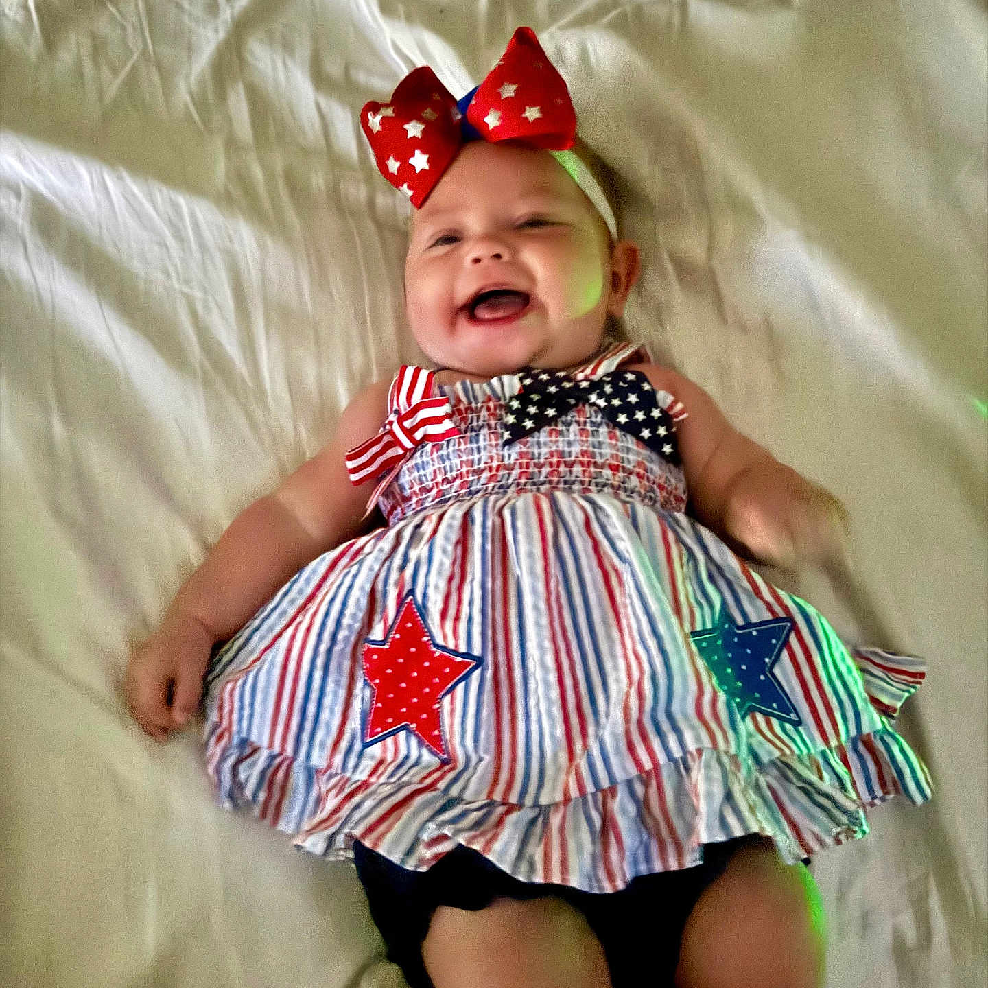 Kennedy is registered to the contest to win money with this photo: baby, child, smiling, lying_down, dress, headband, bow, stars, stripes, red, white, blue, fabric, bed, sheet, happy, cute, infant, person, portrait