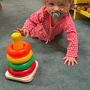 Koura is registered to the contest to win money with this photo: baby, child, crawling, toy, stacking_toy, carpet, pink_clothing, polka_dot, pacifier, indoor, floor, shelf, wooden_toys, yellow_shelf, hand, face, hair, playtime, curious, person