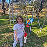 child, playground, swing, climbing_structure, grass, trees, sunny, outdoor, smiling, toy, holiday_hat, casual_clothing, blue_sky, park, nature, person, daylight, happy, footwear, leaves