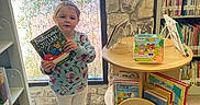 Olivia is registered to the contest to win money with this photo: toddler, child, book, library, bookshelf, window, floor, clothing, shoes, reading, indoor, person, book_display, tile_floor, stone_wall, green_rug, smiling, hair_tie, colorful, young_child