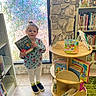 toddler, child, book, library, bookshelf, window, floor, clothing, shoes, reading, indoor, person, book_display, tile_floor, stone_wall, green_rug, smiling, hair_tie, colorful, young_child