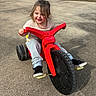 child, toddler, tricycle, red, outdoor, concrete, smiling, casual_clothing, black_shoes, jeans, sunny, playground, happy, vehicle, toy, person, daytime, hair, expression, fun