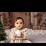 baby, child, christmas_tree, cozy, curious, cute, decorations, dress, festive, holding, holiday, indoor, portrait, seated, smiling, soft_lighting, toddler, white_ball, winter, young_child