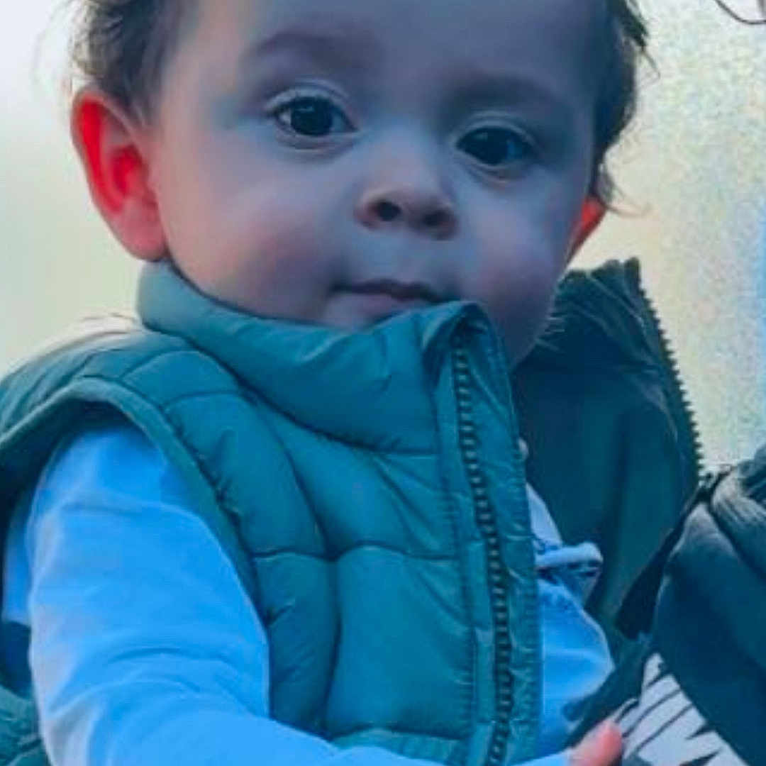 Ayan a rejoint le concours — aidez-le/la à gagner de superbes lots ! adult, blue_shirt, child, clothing, curious, curly_hair, cute, expression, face, green_vest, hand, holding, indoor, light, nike_jacket, person, portrait, soft_background, toddler, young_child