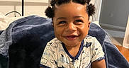 Marcus is registered to the contest to win money with this photo: baby, infant, smile, happy, bantu_knots, hair, onesie, shark_pattern, toy, wooden_beads, bed, blanket, indoor, child, cute, person, skin, sitting, room, joyful