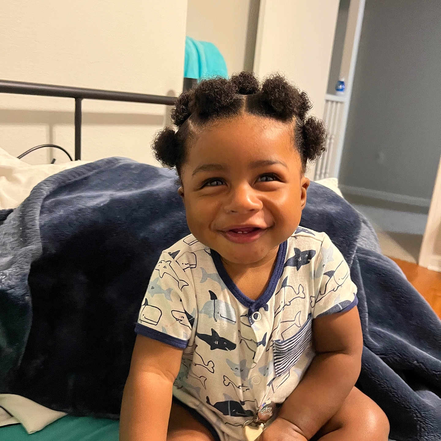 Marcus is registered to the contest to win money with this photo: baby, infant, smile, happy, bantu_knots, hair, onesie, shark_pattern, toy, wooden_beads, bed, blanket, indoor, child, cute, person, skin, sitting, room, joyful