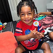 Marcus is registered to the contest to win money with this photo: toddler, child, smiling, clapping, braided_hair, red_shirt, bedroom, toy, blanket, indoor, person, happy, sitting, shorts, furniture, curtains, playful, cute, young_child, casual