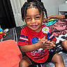 toddler, child, smiling, clapping, braided_hair, red_shirt, bedroom, toy, blanket, indoor, person, happy, sitting, shorts, furniture, curtains, playful, cute, young_child, casual
