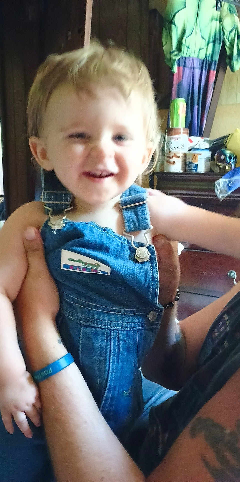 Alastor is registered to the contest to win money with this photo: toddler, child, denim_overalls, smiling, adult_hand, tattoo, wristband, indoor, dresser, clothing, can, furniture, person, holding, happy, face, arm, room, casual, portrait