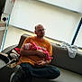 man, baby, newborn, couch, orange_shirt, glasses, indoor, window, blinds, baby_clothes, striped_hat, seated, holding, casual, relaxed, cross-legged, tired, resting, parent, family