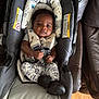 baby, infant, car_seat, seat_harness, clothing, black_shoes, patterned_pants, face, person, indoor, cushion, leather_couch, wood_floor, child, young_child, sitting, serious_expression, baby_boy, baby_girl, child_seat