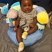 Michael joined the competition — help win amazing prizes! baby, bed, blanket, blue, child, cute, dinosaur_shape, green, happy, indoor, infant, pacifier, person, plush_pillow, seated, smiling, soft_toys, striped_clothing, yellow, yellow_socks