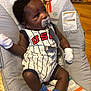 baby, infant, pacifier, onesie, usa, socks, sports, baby_seat, indoor, floor, wooden_floor, clothing, person, cute, child, young, pattern, toy_monkey, smile, white_mittens