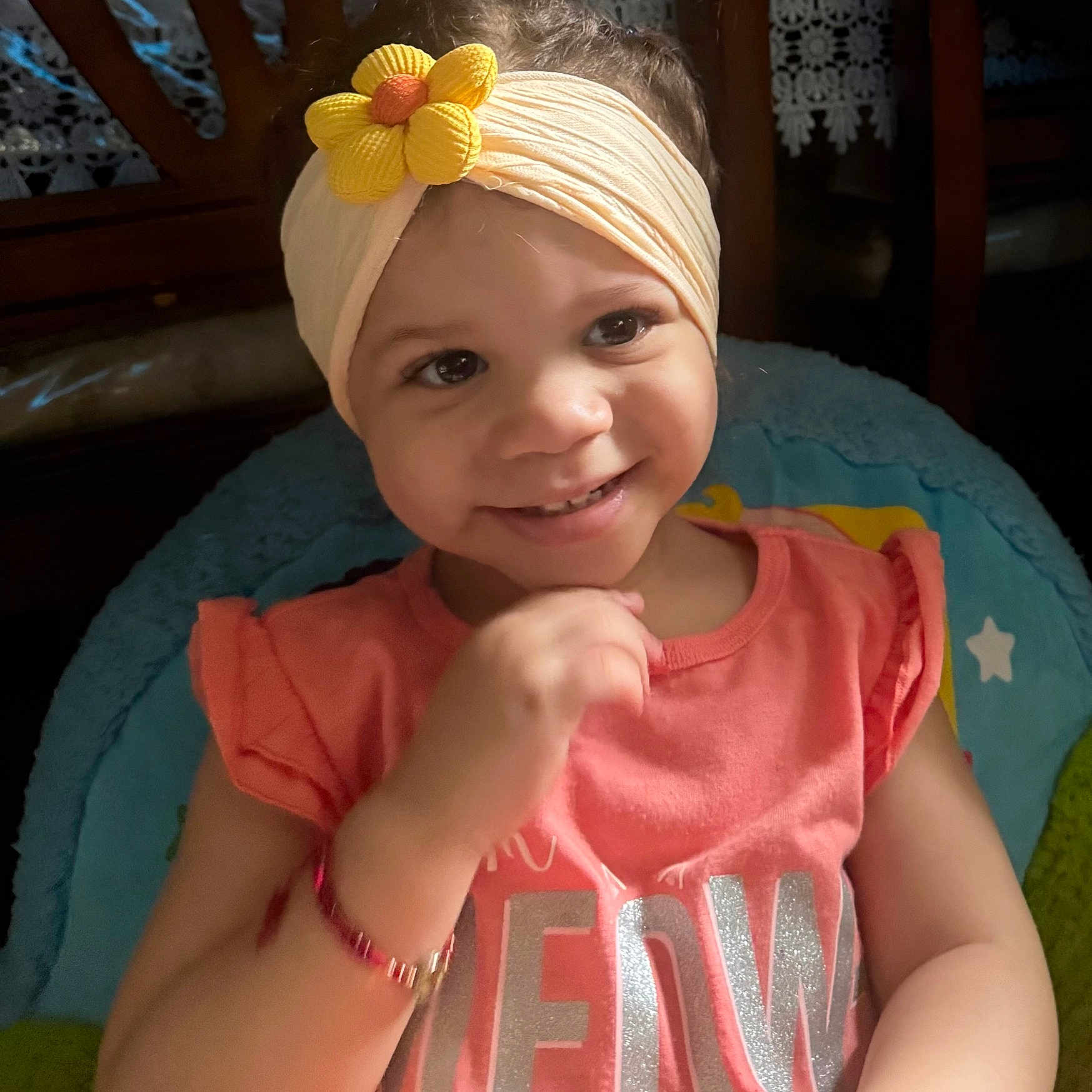 Caitana is registered to the contest to win money with this photo: baby, child, infant, smiling, portrait, close_up, flower_headband, headband, pink_clothing, dress, chubby_cheeks, raised_fist, sitting, baby_seat, blue_cushion, wooden_background, indoor, face, happy, photography