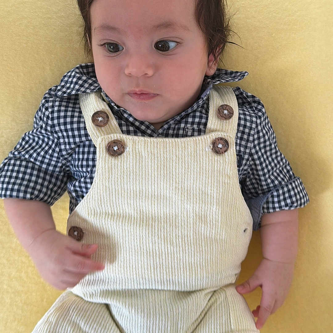 Matheo is registered to the contest to win money with this photo: baby, infant, child, yellow_background, overalls, corduroy, checkered_shirt, wooden_buttons, lying_down, curious_expression, soft_texture, indoors, portrait, young_child, cute, small_hands, short_hair, casual_clothing, comfortable, resting