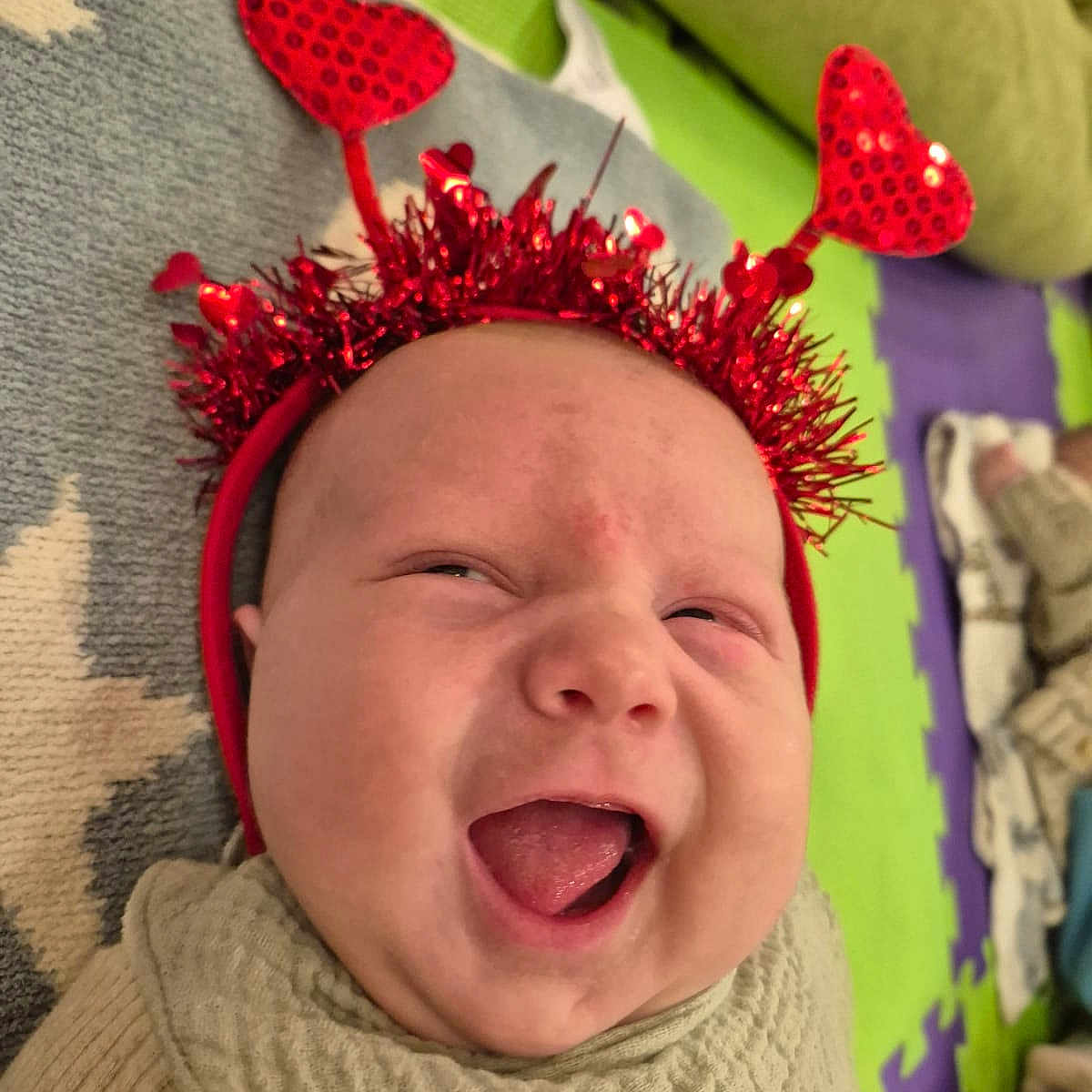 Walter is registered to the contest to win money with this photo: antennae, baby, beige, child, closeup, clothing, expression, face, green, headband, heart, indoors, infant, laughing, pattern, play_mat, purple, red, smiling, star