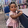 toddler, child, girl, pink_dress, standing, library, bookshelf, chair, carpet, curly_hair, hair_ties, footwear, indoor, young_child, person, furniture, expression, curiosity, portrait, casual_clothing