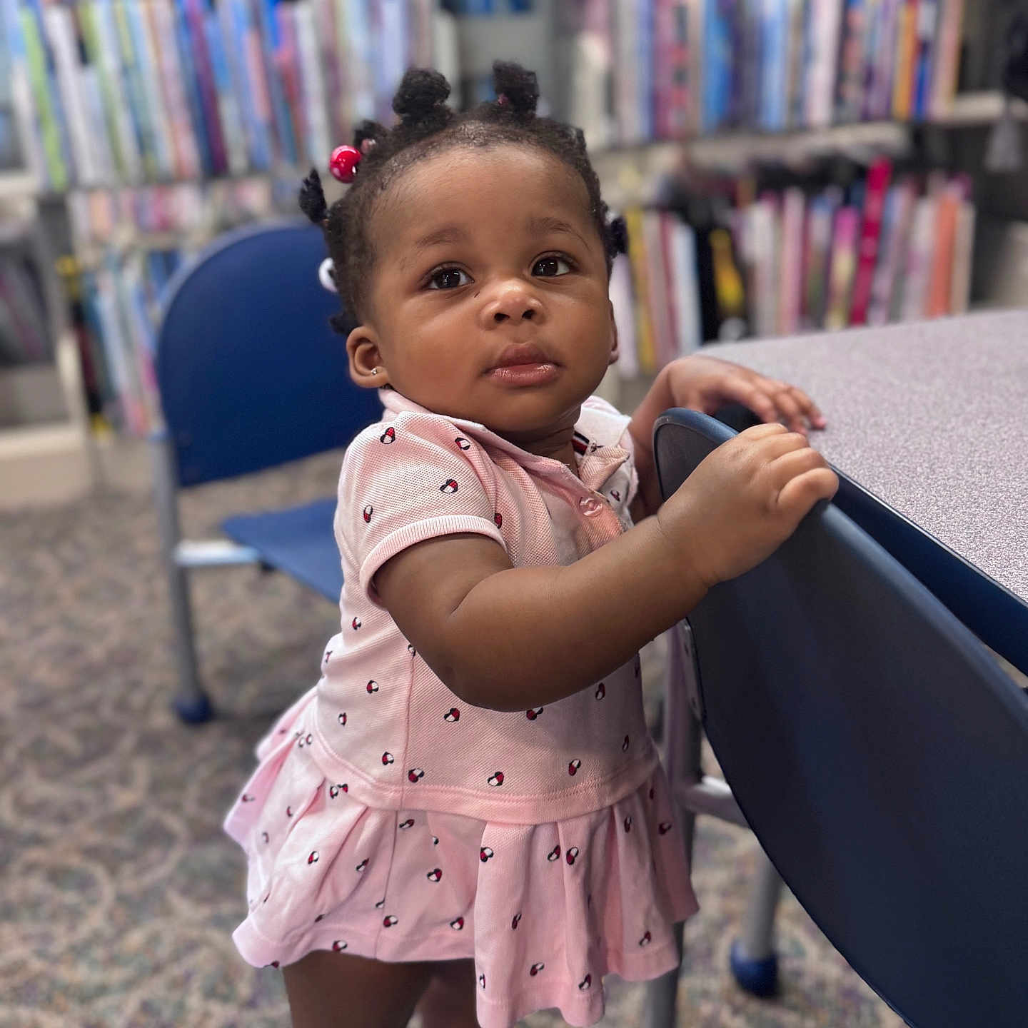 Ryanne joined the competition — help win amazing prizes! toddler, child, girl, pink_dress, standing, library, bookshelf, chair, carpet, curly_hair, hair_ties, footwear, indoor, young_child, person, furniture, expression, curiosity, portrait, casual_clothing