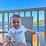 baby, child, smiling, white_shirt, striped_shorts, chair, wicker, balcony, sea_view, mountains, outdoor, daylight, blue_sky, railing, barefoot, cute, portrait, infant, summer, relaxing
