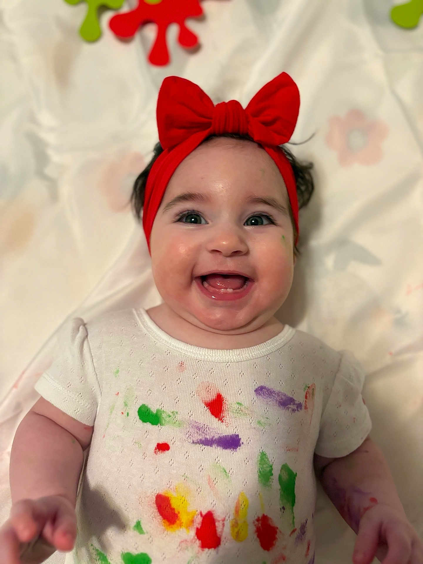 Salmah Pérez Acosta joined the competition — help win amazing prizes! baby, child, smiling, red_bow, headband, paint_splatter, white_shirt, happy, face, arm, skin, indoors, closeup, person, infant, cute, cheerful, expression, colorful, playful