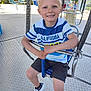 amusement_park, blond_hair, blue_seat, boy, casual_clothing, child, daylight, fun, happy, holding, metal_bars, outdoor, playground, recreation, shorts, sitting, smiling, swing, tshirt, white_shoes