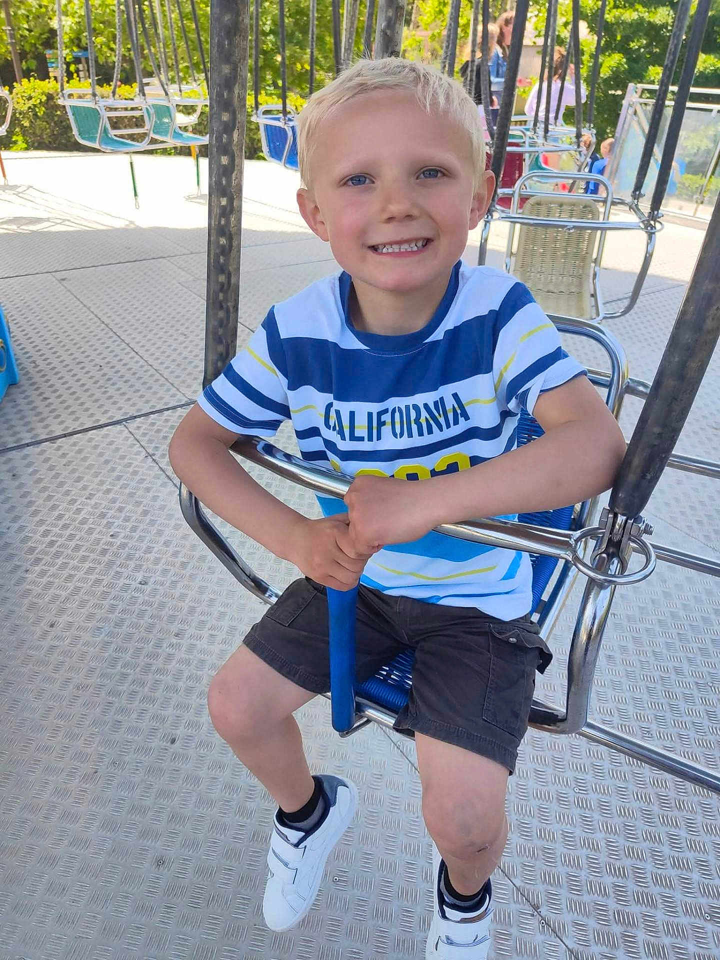 Lucas a rejoint le concours — aidez-le/la à gagner de superbes lots ! child, boy, smiling, sitting, swing, amusement_park, outdoor, daylight, shorts, tshirt, white_shoes, blue_seat, playground, happy, metal_bars, blond_hair, casual_clothing, holding, fun, recreation