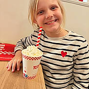 Emma a rejoint le concours — aidez-le/la à gagner de superbes lots ! beverage, blonde_hair, casual_clothing, child, cup, drink, food, girl, happy, indoor, person, railing, red_heart, seated, smiling, straw, striped_shirt, whipped_cream, window, wooden_table