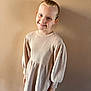 Emma a rejoint le concours — aidez-le/la à gagner de superbes lots ! beige_dress, blue_eyes, casual_clothing, child, cozy, cute, fashion, girl, happy, indoor, knitwear, light_skin, long_sleeves, portrait, smile, standing, wall, warm_colors, young_person, youth