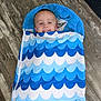 child, smiling, sleeping_bag, blue, waves, shark, floor, wooden_floor, indoors, cozy, happy, face, person, zipper, pattern, head, toy, home, carpet, blanket
