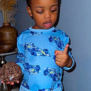 Saidou Bah joined the competition — help win amazing prizes! child, toddler, pajamas, blue_clothing, indoor, decor, vase, wicker_ball, wood_floor, counting, curious, standing, shorts, patterned_clothing, person, face, hand, furniture, wall, home