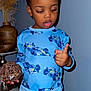 child, toddler, pajamas, blue_clothing, indoor, decor, vase, wicker_ball, wood_floor, counting, curious, standing, shorts, patterned_clothing, person, face, hand, furniture, wall, home