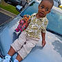 Saidou Bah is registered to the contest to win money with this photo: child, toy, car, parking_lot, sneakers, shirt, shorts, tree, building, reflection, side_view, outdoor, daytime, curious, person, vehicle, colorful, casual_clothing, holding, sitting